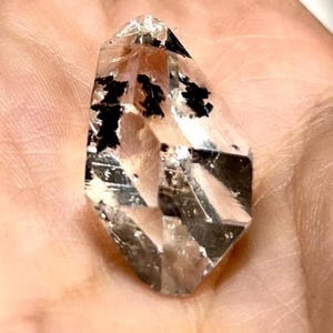 Peut inclure: Un cristal de quartz transparent et facetté présentant des inclusions noires distinctes et irrégulières dispersées à l'intérieur. La pierre a une forme pointue et allongée avec de multiples surfaces lisses et réfléchissantes.