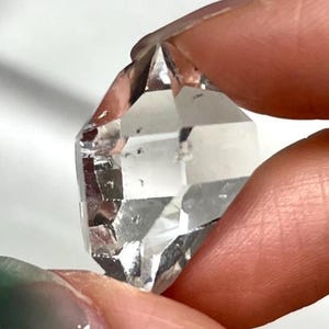 Puede incluir: Un cristal de cuarzo transparente de doble terminación con forma hexagonal. La piedra presenta bordes geométricos afilados y una apariencia transparente similar al vidrio con inclusiones internas visibles y reflejos de luz naturales.