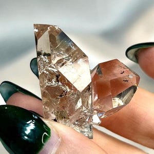 Peut inclure: Un petit amas de cristaux de quartz translucides à double terminaison, présentant une teinte brun clair fumé. Le spécimen minéral possède des facettes géométriques nettes et des inclusions internes visibles, avec une texture vitreuse.