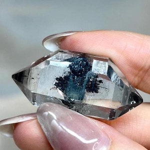 Puede incluir: Un cristal de cuarzo transparente de doble terminación que presenta una prominente inclusión mineral de color azul oscuro a negro en su centro. El cristal tiene una estructura facetada y transparente con extremos puntiagudos y superficie lisa.