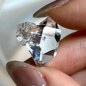 Peut inclure: Un cristal de quartz transparent à facettes avec un amas texturé et rugueux attaché sur un côté. La pierre présente de multiples surfaces polies qui reflètent la lumière, révélant un aspect transparent avec des inclusions internes et des bords nets.
