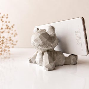 Könnte beinhalten: Ein grauer Telefonhalter in Bärenform aus Beton mit geometrischem Design. Der Bär hält ein silbernes Smartphone. Der Hintergrund ist neutralfarben mit einem Zweig getrockneter Blumen.