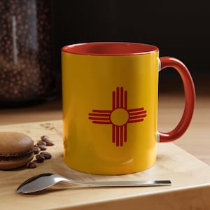 May include: A yellow coffee mug with a red handle and interior. The mug features the New Mexico state flag, a red Zia symbol, centered on the yellow surface. A spoon and a macaron are on a wooden surface.