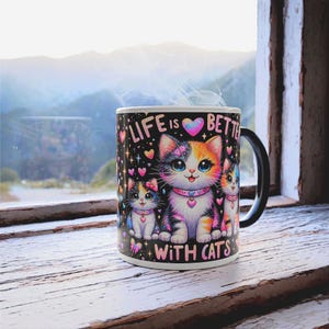 May include: A black ceramic mug featuring the phrase "LIFE IS BETTER WITH CATS" and images of three cartoon cats. The cats are depicted in blue, orange, and white, with pink heart details. The mug has a black handle and is on a white wooden surface.
