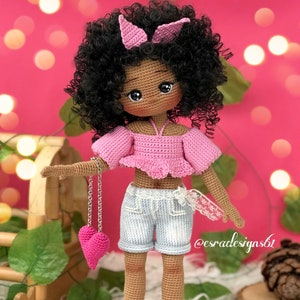 May include: Crocheted doll with brown skin, black curly hair, and a pink top, blue denim shorts, and pink shoes. The doll is holding a pink heart charm on a silver chain.