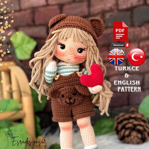 Op de afbeelding: Haakpatroon voor een amigurumipop in het Engels en Turks. De pop draagt een bruin-wit gestreept shirt, een bruine overall met een berenkop, een bruine muts met berenoren en bruine laarzen. De pop houdt een rood hart vast. Het patroon is beschikbaar als PDF-download.