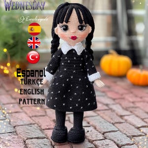 May include: Crocheted doll with black hair, wearing a black dress with white polka dots, white collar, and black shoes. The doll is standing on a brick surface. The text "WEDNESDAY" is visible in the background.