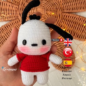 Puede incluir: Un perro de peluche tejido a crochet con orejas negras, una camisa roja y un cuerpo blanco. El juguete está siendo sostenido en una mano. El fondo es una cesta de mimbre.