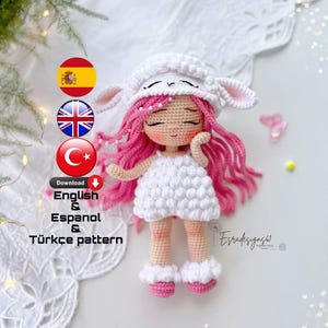 May include: A crocheted amigurumi doll wearing a white sheep costume, pink hair, and pink shoes. The doll is holding its hands together in front of its chest. The image includes text that says "Download English & Espanol & Türkçe pattern".