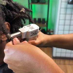 May include: A person using a silver Instant Loc Machine to create dreadlocks in a person's hair. The machine has the words "Instant Loc Machine" printed on it.