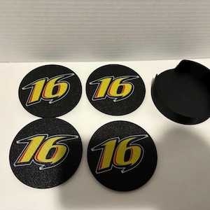 May include: A set of four black, round coasters featuring the number "16" in yellow and red, and a matching black holder. The coasters have a textured surface.