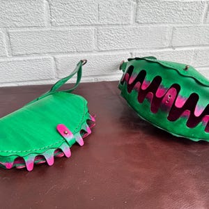 May include: Two green leather bags with pink and green accents. One bag is a clutch with a handle, and the other is a larger, open-top bag. Both bags have a unique design with a wavy, cut-out pattern.