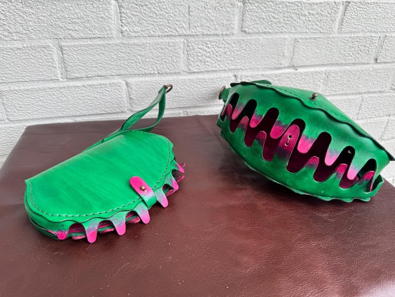 May include: Two green leather bags with pink and green accents. One bag is a clutch with a handle, and the other is a larger, open-top bag. Both bags have a unique design with a wavy, cut-out pattern.