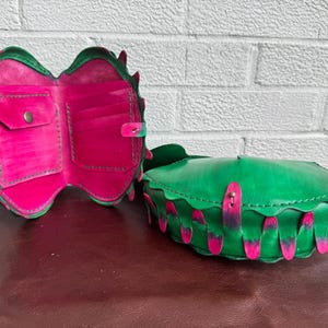 May include: A green leather wallet and matching handbag with a unique design. The wallet is open, revealing pink interior pockets. The handbag is closed, with pink accents. Both items have a scalloped edge.