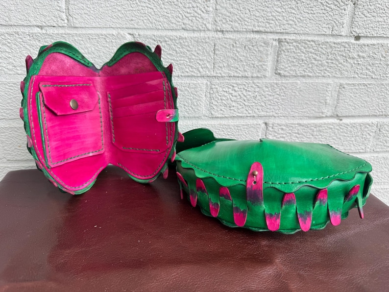 May include: A green leather wallet and matching handbag with a unique design. The wallet is open, revealing pink interior pockets. The handbag is closed, with pink accents. Both items have a scalloped edge.