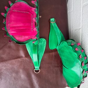 May include: A handmade leather bag and strap set. The bag is a vibrant pink with green trim and a decorative edge. The strap is green with a metal ring. The set is displayed on a brown leather surface.