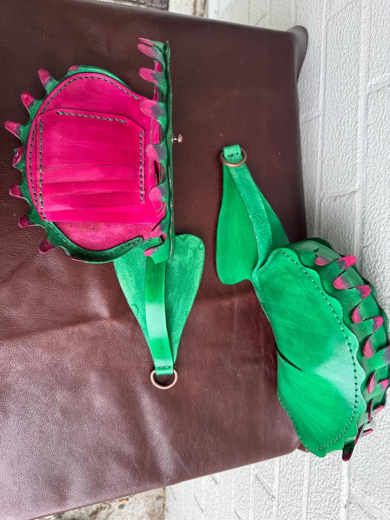May include: A handmade leather bag and strap set. The bag is a vibrant pink with green trim and a decorative edge. The strap is green with a metal ring. The set is displayed on a brown leather surface.
