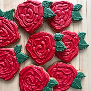 Puede incluir: Nueve galletas rojas con forma de rosa con detalles de hojas verdes están dispuestas sobre una superficie de madera clara. Las galletas tienen un glaseado detallado, que se asemeja a pétalos de rosa. Las hojas verdes añaden contraste.