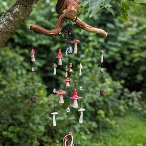 Pode incluir: Um mobile caprichoso com ornamentos de cogumelos em miniatura com chapéus vermelhos e caules brancos, suspensos de um galho de madeira natural. Contas de cristal transparente e pequenas pinhas completam o design. Pendurado ao ar livre.