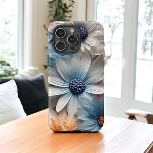 May include: A phone case with a floral design featuring white and blue paper flowers. The case has a black camera bump with three lenses.