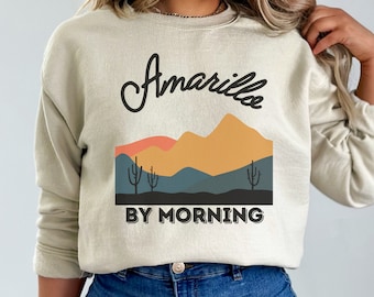 May include: A beige sweatshirt with a graphic design featuring the text "Amarillo by Morning" and a stylized mountain range with a sunset.