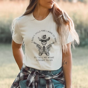 以下が含まれることがあります： 白地に黒と白のグラフィックがプリントされたTシャツ。カウボーイハットをかぶり、2丁のピストルを持った骸骨が描かれている。Tシャツには「I don't need the laws of man to tell me what I ought to do.」という文字が書かれている