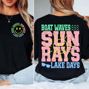 Op de afbeelding: Zwart T-shirt met een grafische print van een lachende zon met de tekst "Boat Waves Sun Rays Lake Days" in kleurrijke letters.
