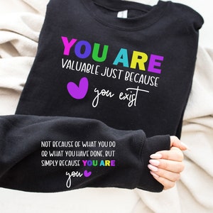 May include: Black sweatshirt with a rainbow colored text that reads "YOU ARE VALUABLE JUST BECAUSE you exist" and a purple heart. The text continues on the sleeve, "NOT BECAUSE OF WHAT YOU DO OR WHAT YOU HAVE DONE. BUT SIMPLY BECAUSE YOU ARE you" with a small pink heart.