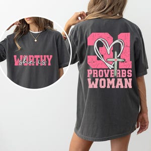 May include: A gray t-shirt with a pink graphic on the back that says "Proverbs 31 Woman" with a heart and cross. The front of the shirt has a pink graphic that says "She is Worthy".