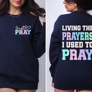 May include: Navy blue sweatshirt with a rainbow colored graphic on the front that says "Just Pray" and a rainbow colored graphic on the back that says "Living the Prayers I Used to Pray".