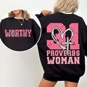 May include: Black sweatshirt with pink text on the front that says "she is WORTHY" and pink text on the back that says "PROVERBS WOMAN" with a pink heart and cross graphic.