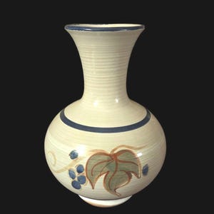 May include: Cream-colored ceramic vase with a round body and a flared neck. The vase features a blue rim and a blue band around the body. A painted design of grapes and leaves in shades of blue, green, and brown decorates the front.