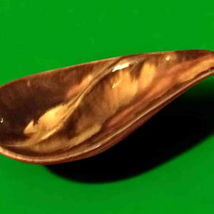 May include: A brown and tan ceramic spoon rest with a curved handle. The spoon rest has a glossy finish and a unique, organic shape. The interior features a marbled pattern of light and dark brown tones. The background is a solid green.