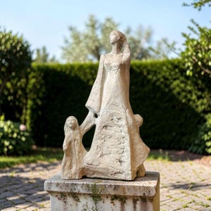 May include: A beige stone sculpture depicting a tall central figure standing with arms raised, flanked by two smaller figures on either side. The textured surface shows abstract, carved details and flowing lines throughout the entire composition.
