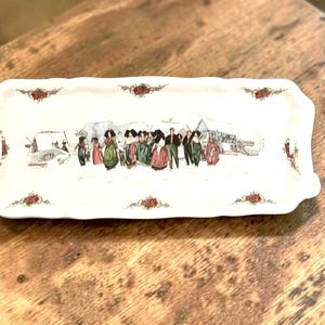 May include: A rectangular white ceramic tray with scalloped edges, featuring a central illustration of a group of people in colorful clothing walking through a snowy landscape, accented by small floral motifs along the rim of the dish.