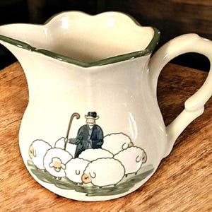 May include: Cream-colored ceramic pitcher with a scalloped rim and a curved handle. The pitcher features a pastoral scene of a shepherd with a cane and a flock of sheep. The rim is trimmed with green paint.