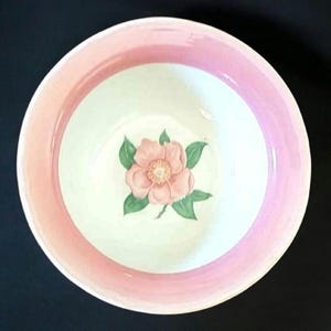 May include: A round ceramic bowl with a pink rim and a white interior. The bowl features a pink flower with green leaves in the center. The bowl is likely used for serving food or as a decorative piece.