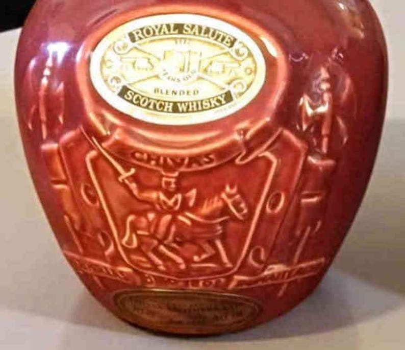 K&ouml;nnte beinhalten: Eine rote Keramikflasche Royal Salute Blended Scotch Whisky. Die Flasche hat ein goldenes Etikett und gepr&auml;gte Details, darunter einen Ritter auf einem Pferd. Der Text "Royal Salute" und "Blended Scotch Whisky" sind sichtbar.