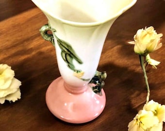 May include: A vintage ceramic vase with a pink base and a white upper section. The vase features green leaf and yellow flower accents. The vase has a flared rim and two small handles. The vase is sitting on a wooden surface.