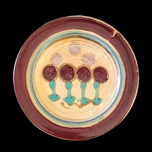 May include: A round, ceramic plate with a maroon rim and a light yellow center. The center features four maroon circles on turquoise stems. A light blue ring separates the rim from the center. The plate has a rustic, handmade appearance.