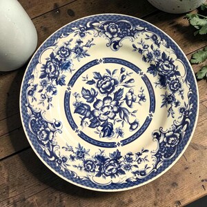 May include: A round, cream-colored plate with a blue floral design. The plate features a central floral arrangement and a decorative border with additional floral motifs. The plate is set on a wooden surface.
