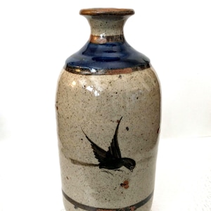 May include: A ceramic vase with a speckled beige body, a dark blue neck, and a brown rim. A black bird is painted on the front. The vase has a cylindrical shape with a slightly flared top.