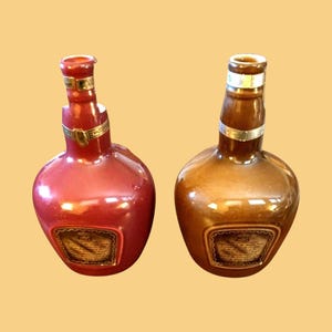 K&ouml;nnte beinhalten: Zwei dekorative Keramikflaschen, eine rote und eine braune, mit rundem Boden und schlankem Hals. Jede Flasche hat ein goldfarbenes Band um den Hals und ein Etikett auf der Vorderseite. Die Flaschen werden vor einem hellgelben Hintergrund pr&auml;sentiert.
