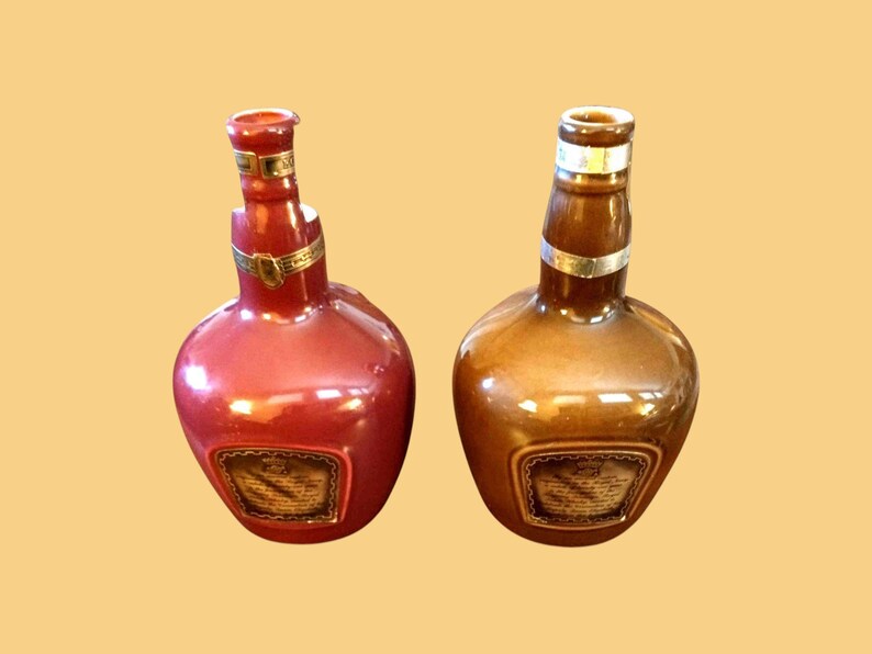 K&ouml;nnte beinhalten: Zwei dekorative Keramikflaschen, eine rote und eine braune, mit rundem Boden und schlankem Hals. Jede Flasche hat ein goldfarbenes Band um den Hals und ein Etikett auf der Vorderseite. Die Flaschen werden vor einem hellgelben Hintergrund pr&auml;sentiert.