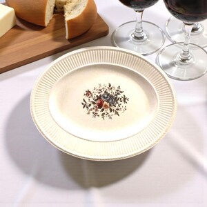 May include: Off-white oval serving platter with a floral design in the center. The platter has a ribbed border and is surrounded by wine glasses, cheese, and bread. The floral design includes red, blue, and tan flowers.