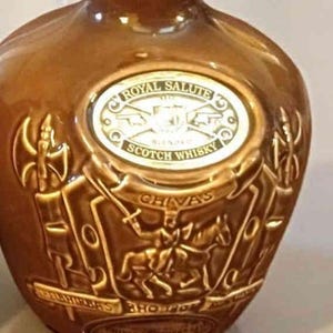 K&ouml;nnte beinhalten: Eine braune Keramikflasche mit gepr&auml;gten goldenen Buchstaben und einem dekorativen Design. Die Flasche tr&auml;gt die Aufschriften "Royal Salute" und "Scotch Whisky". Das Design umfasst einen Ritter zu Pferd und das Wort "Chivas".