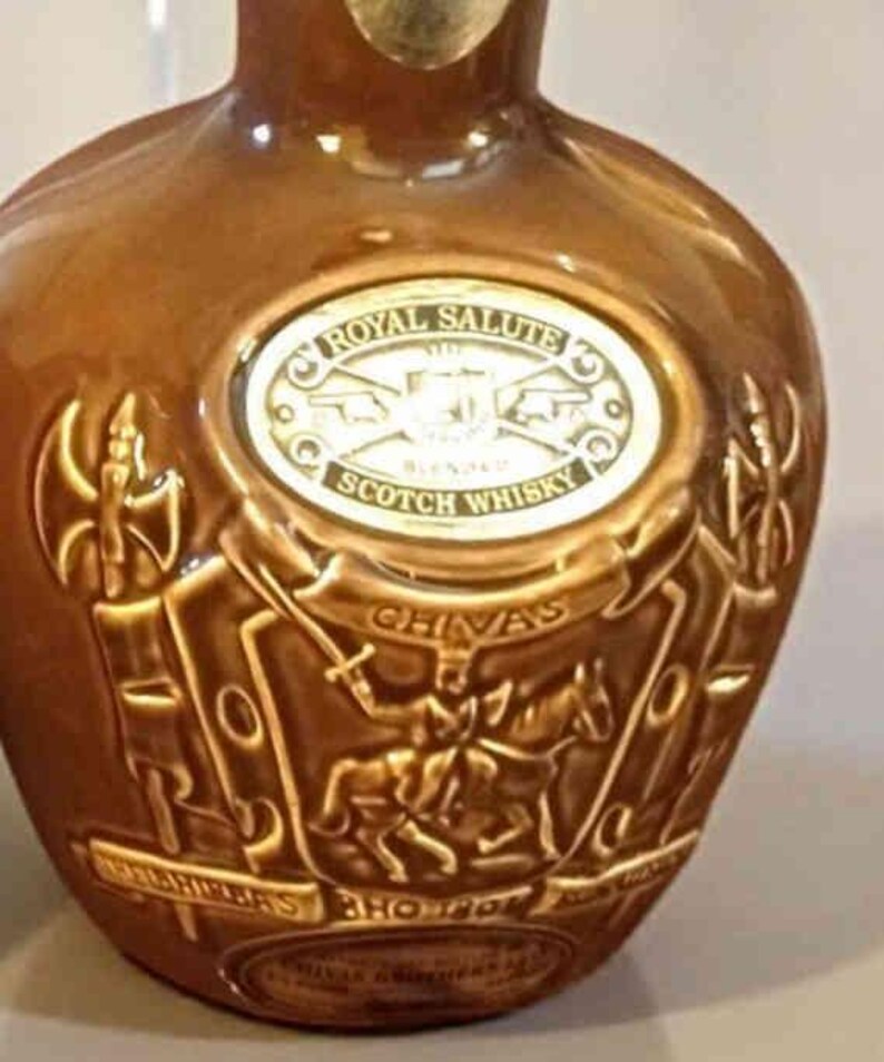 K&ouml;nnte beinhalten: Eine braune Keramikflasche mit gepr&auml;gten goldenen Buchstaben und einem dekorativen Design. Die Flasche tr&auml;gt die Aufschriften "Royal Salute" und "Scotch Whisky". Das Design umfasst einen Ritter zu Pferd und das Wort "Chivas".