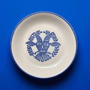 May include: A round, off-white ceramic plate with a blue rim and a blue illustration. The illustration features two birds facing each other, surrounded by a wreath. The plate is set against a blue background.