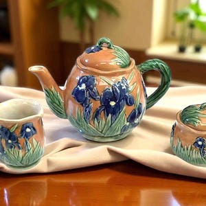 May include: A ceramic tea set featuring a teapot, a sugar bowl, and a mug. The set has a peach-colored base with blue iris flower designs and green accents. The teapot has a curved spout and handle. The sugar bowl has a lid.