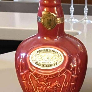 K&ouml;nnte beinhalten: Eine rote Keramikflasche Royal Salute Blended Scotch Whisky. Die Flasche hat goldene Akzente und ein goldenes Etikett mit dem Markennamen. Die Flasche hat eine einzigartige Form und ist mit gepr&auml;gten Bildern von Reitern verziert.